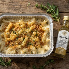 A baked vegan mac and cheese in a square aluminum tray, topped with melted cheese and golden breadcrumbs, shown on a rustic wooden surface with a small bottle of Sabatina olive oil and rosemary sprigs.