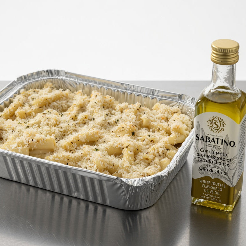 A foil-tray prepared vegan mac and cheese dish with a golden breadcrumb topping sits on a metallic surface next to a bottle of Sabatino white truffle flavored olive oil.