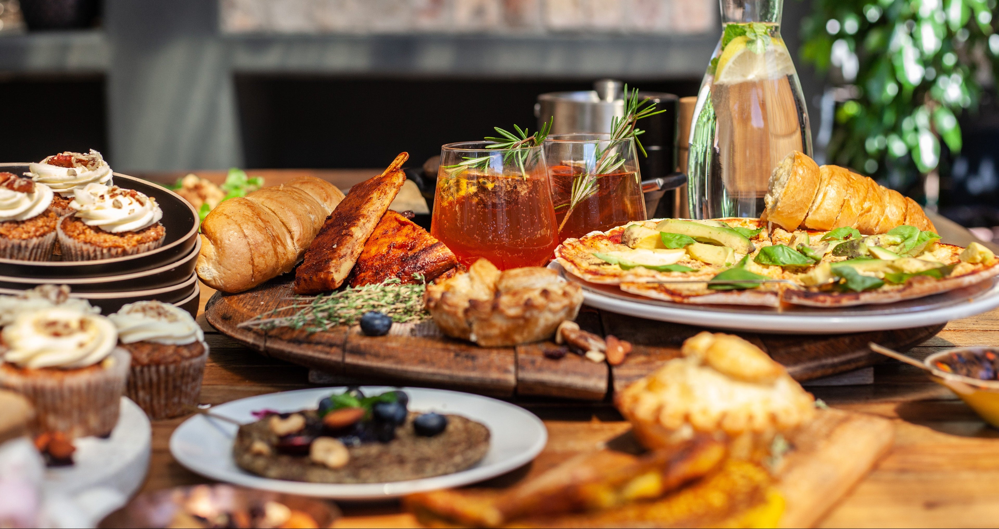 Table with various healthy plant-based foods items including vegan margherita pizza, garlic bread, vegan carrot cupcakes and vegan chicken mushroom pies with a glass of water.