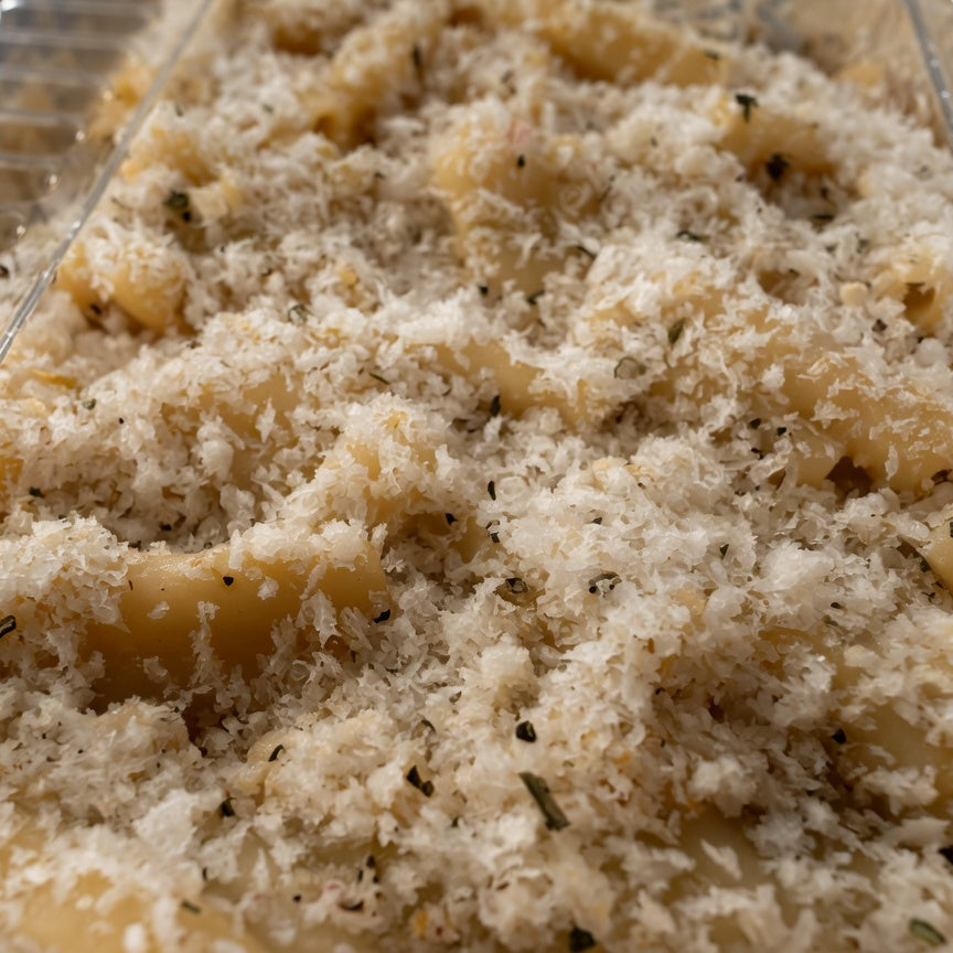 Close-up of a baked vegan mac and cheese dish topped with a thick layer of melted cheese and breadcrumbs, served in a clear plastic baking dish.
