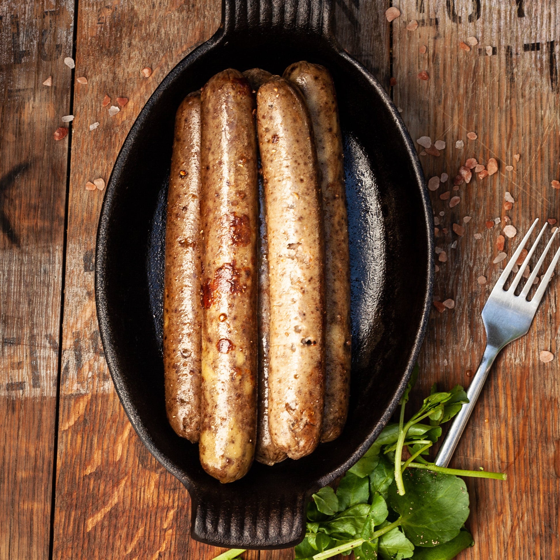 Three sausages in a black cast iron skillet on a wooden surface with a fork and greens.
