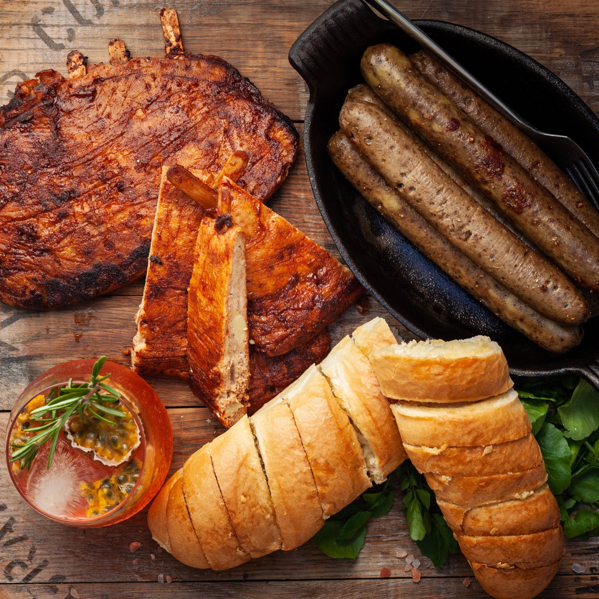 Vegan meat platter with roasted vegan meat, vegan boerewors plant-based sausage, garlic bread, and a drink on a wooden surface.