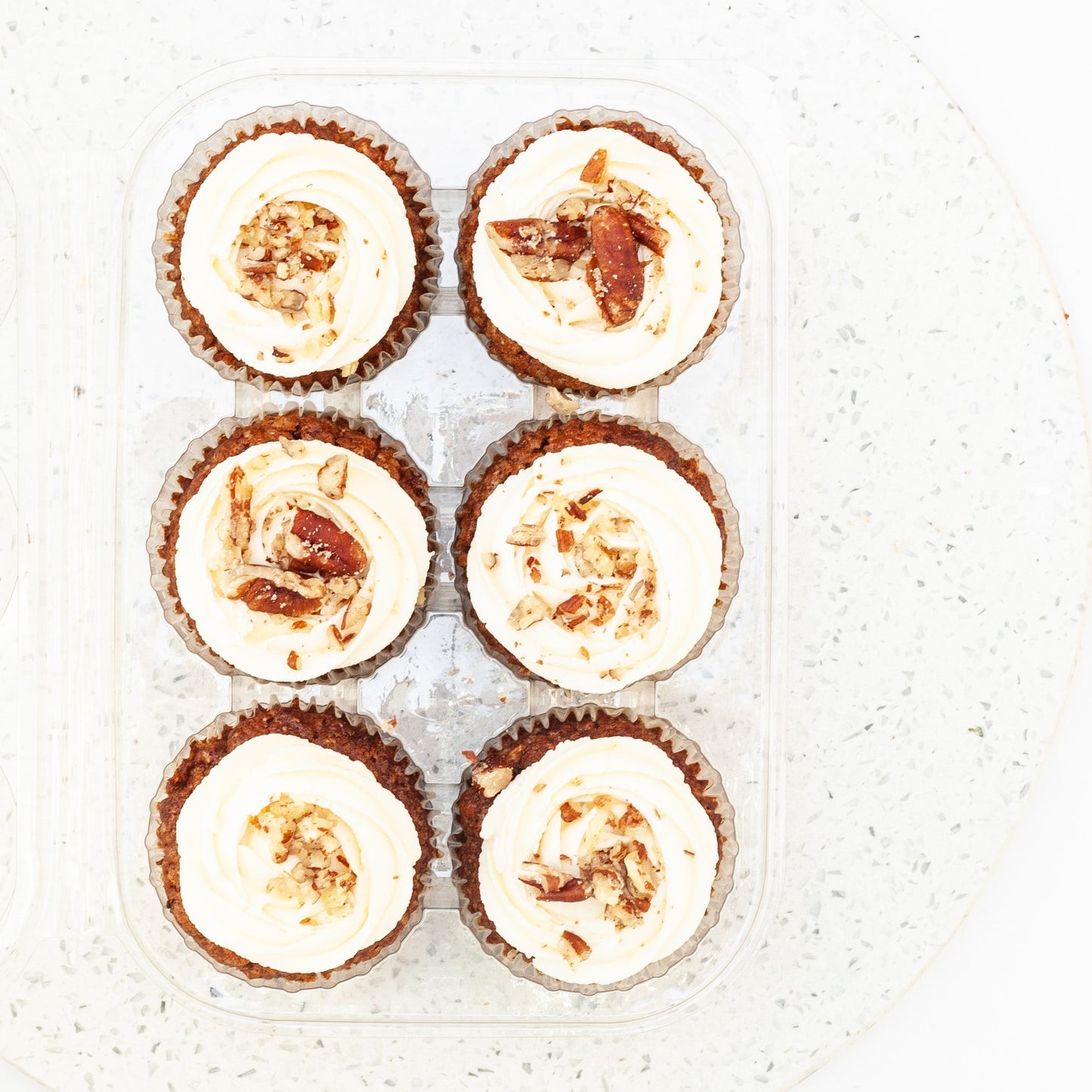 Six vegan carrot cupcakes with white frosting and crushed nuts on a textured white surface.