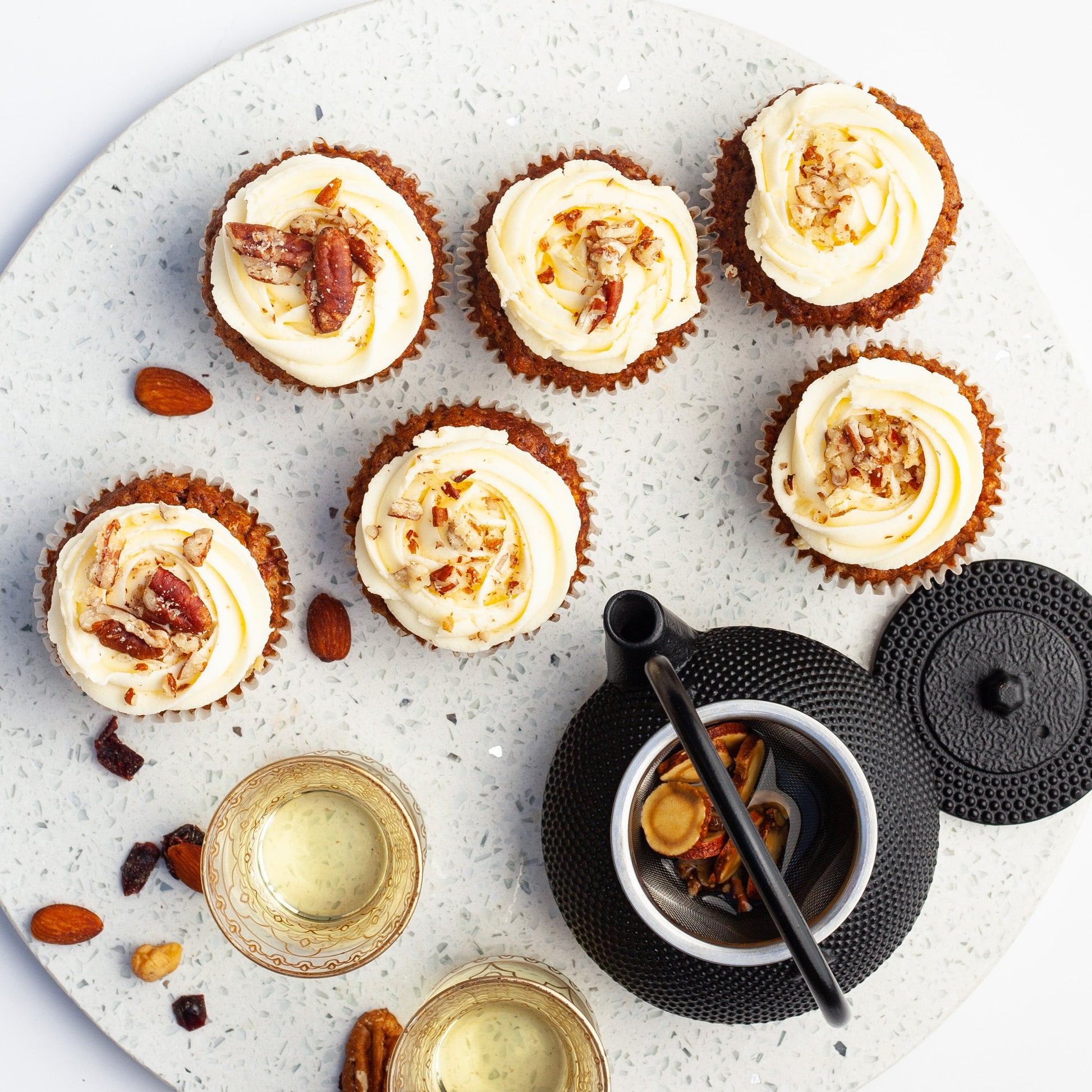 Vegan carrot cupcakes with white frosting on a round stone board with herbal tea and nuts.