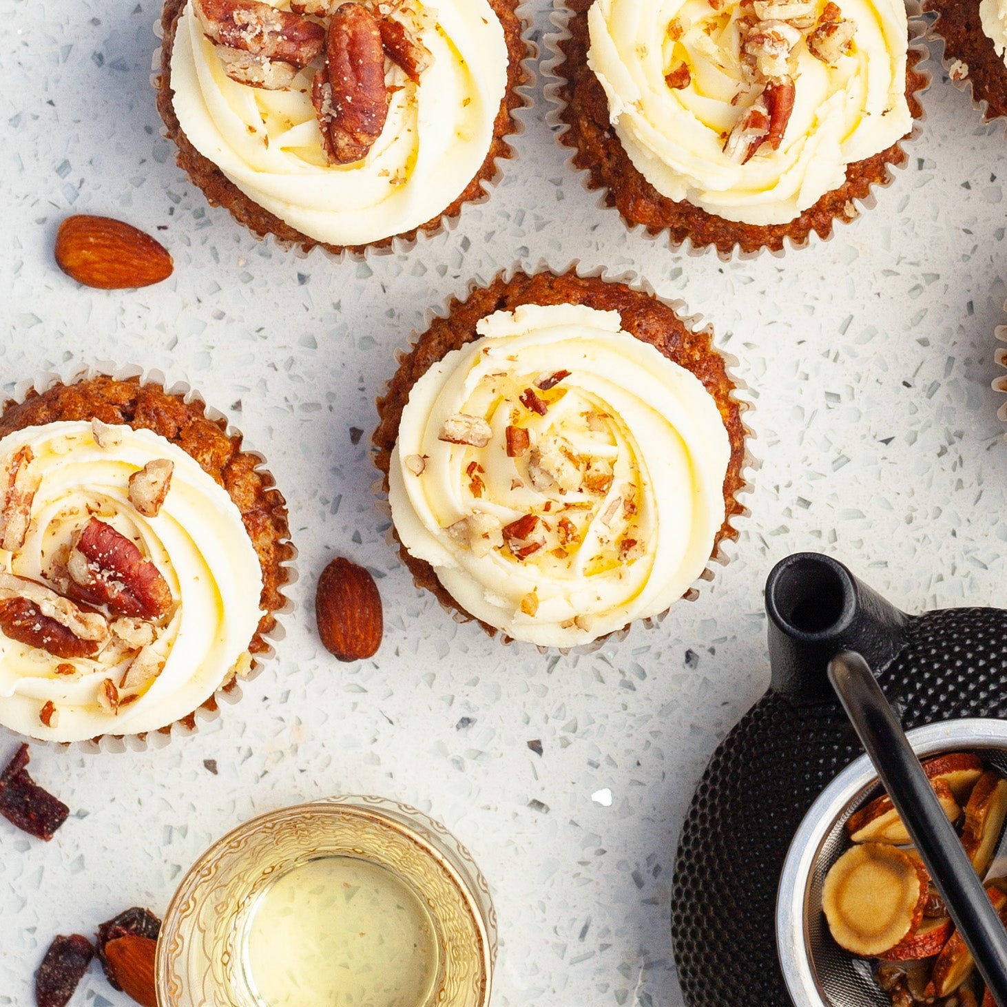 Vegan carrot cupcakes with white frosting and pecan nuts on a textured surface with herbal teas. 