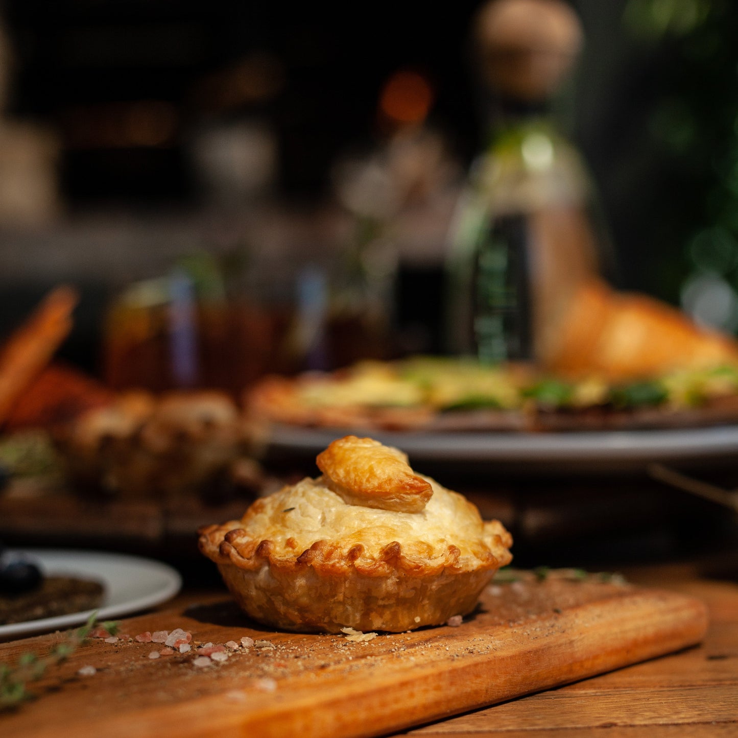 A vegan chicken mushroom pie on a wooden board with a blurred background of other vegan food items and bottles.