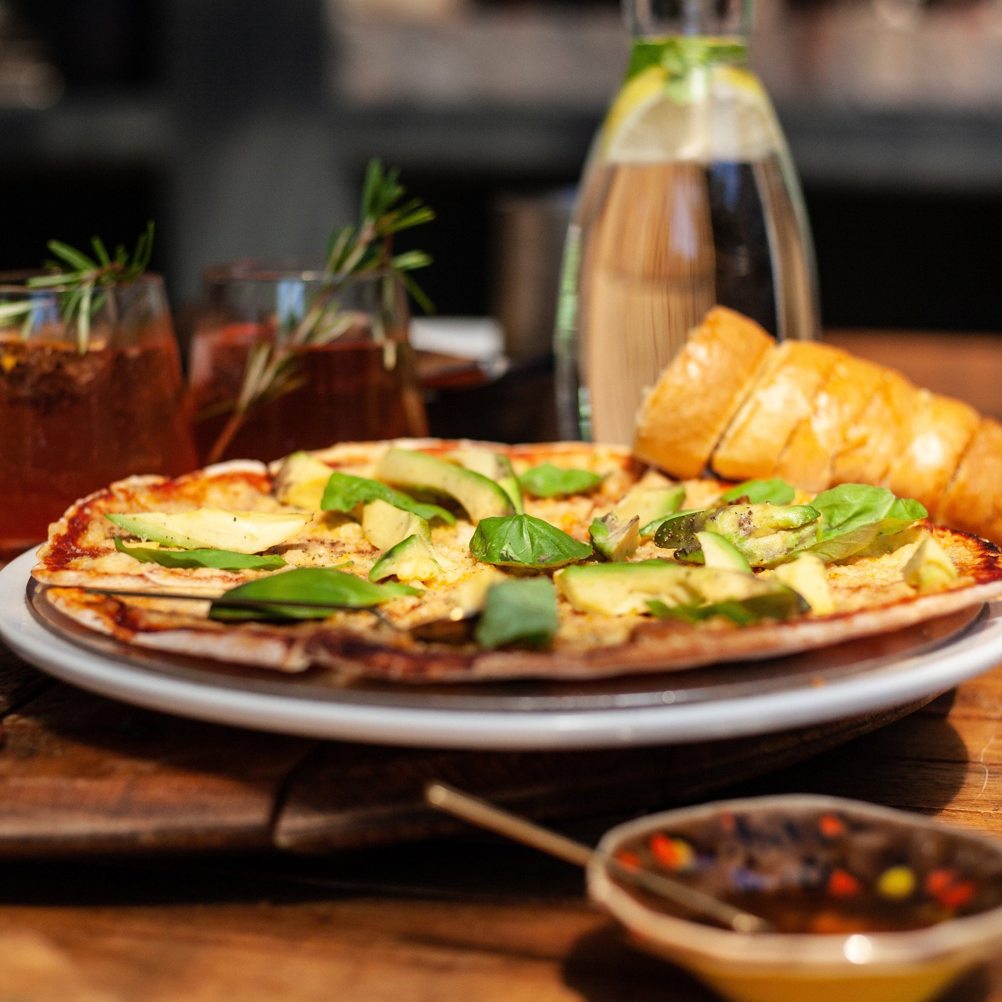 vegan margherita pizza with avocado on a wooden table with drinks and a bottle in the background